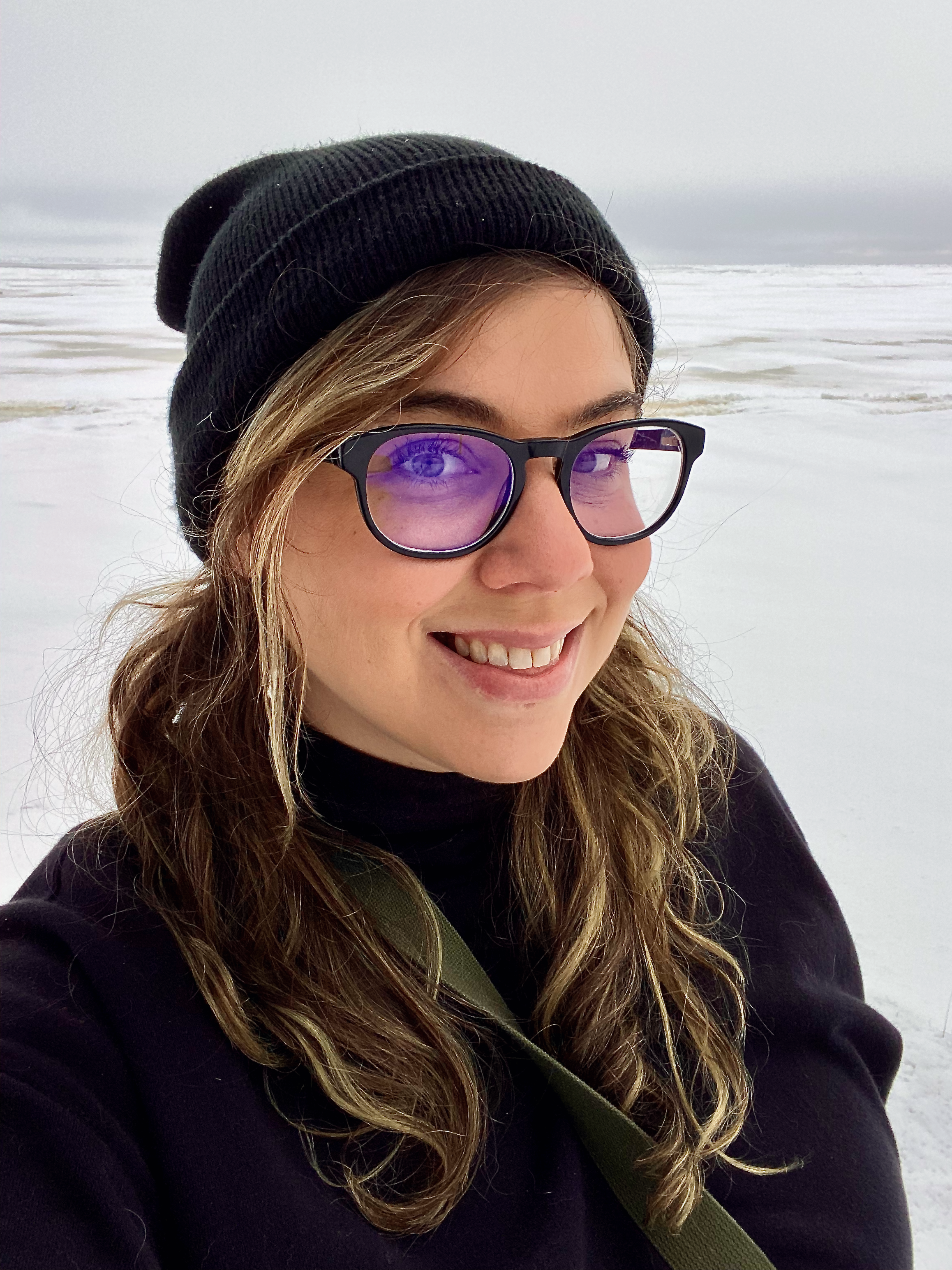femme presenting human with brown hair and blond highlights, black thick framed glasses and black jacket and hoodie with frozen Lake Superior and blue grey sky in background